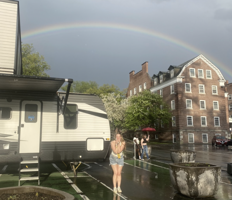 Alt text: A student stands excitedly in front of a trailer as a vivid rainbow arcs across the sky behind Dartmouth dorm buildings.