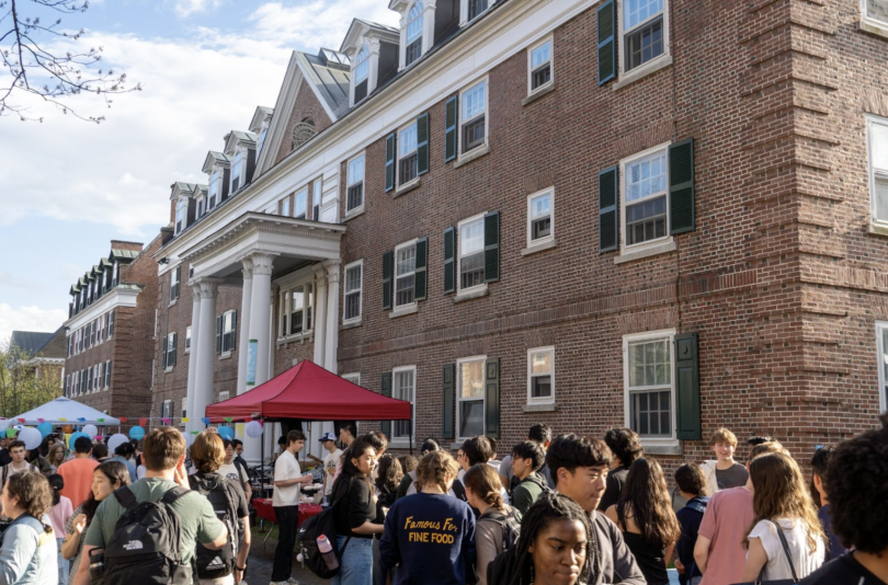 Students crowding Massachusetts Row, waiting in lines for food. Red and white tents poke out behind the crowd. In the background is Mid-Mass Hall, a rectangular, red brick building with white window edging and green shutters.