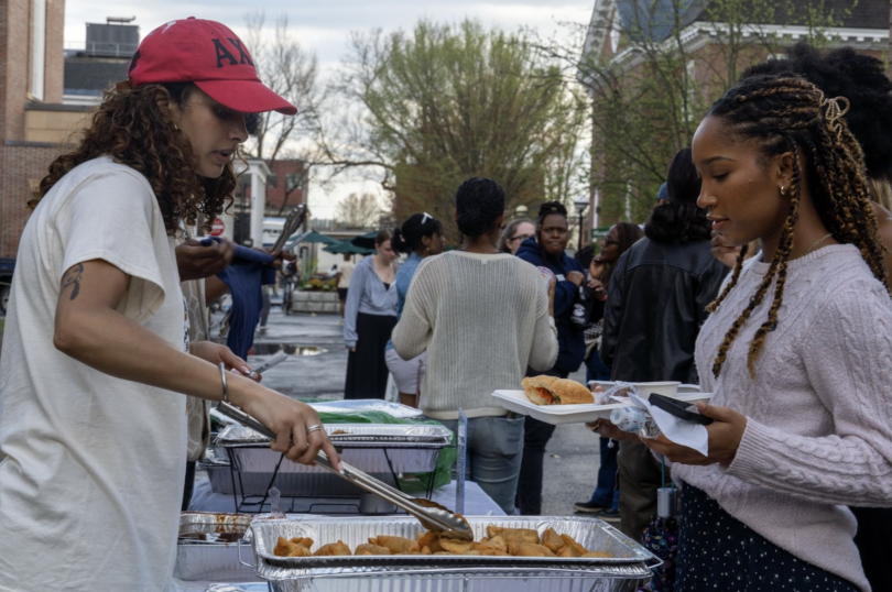 A student using tongs to serve a samosa to another student holding out a tray. In the distance is a crowd of people.