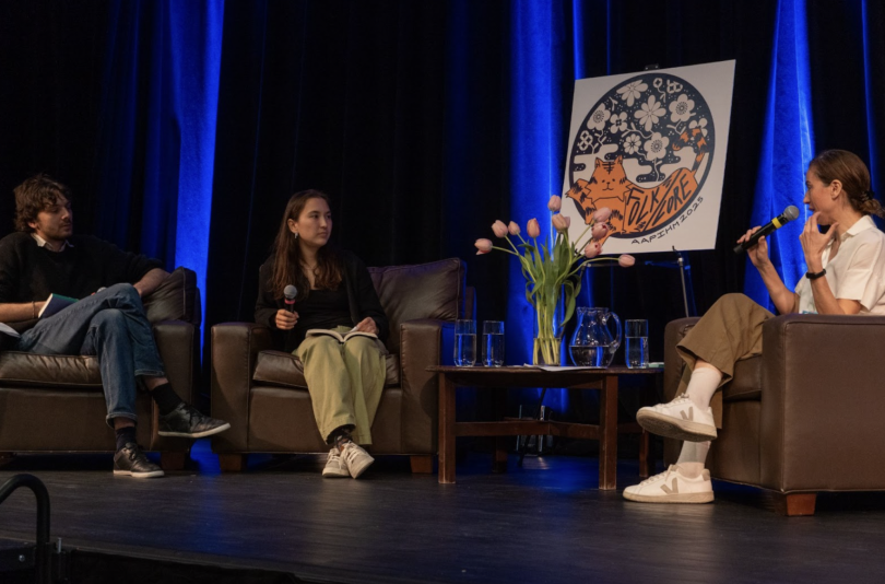 Three people sit in brown armchairs, two to the left and one to the right. The person to the right, Zaina Arafat, is speaking into a microphone. In the middle is a short table with a vase of pink tulips and glasses of water. In the background is