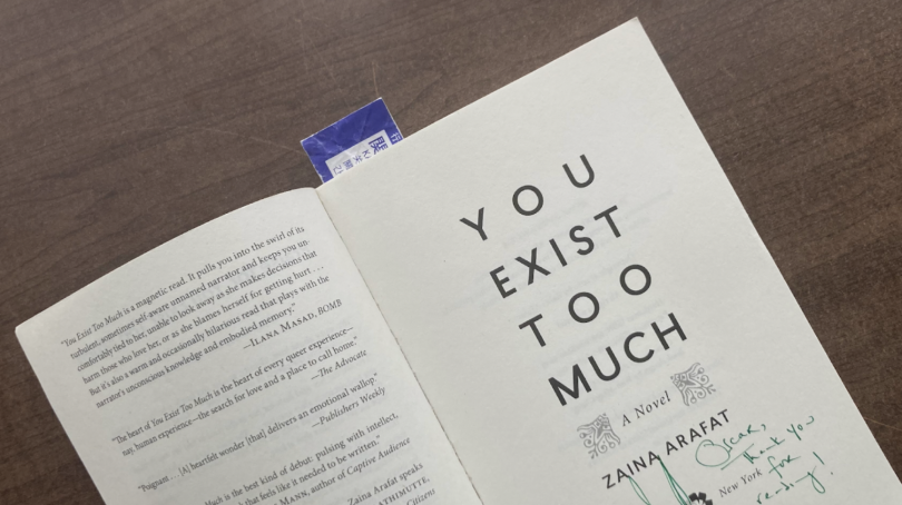 My signed copy of You Exist Too Much, against a dark brown wooden background. A note is written in green pen on the title page.