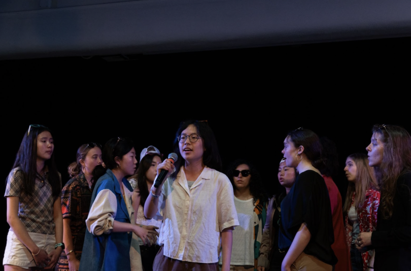 A group of students sing, forming a circle around a student in the center who is also singing into a microphone.