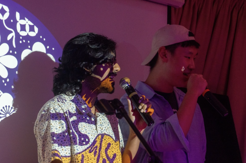 Two students singing a song, both are standing to the side, facing right and holding microphones. The AAPIHM logo is projected on top of one of the students.