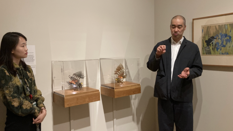 Choe U-Ram, a tall man in a white button-down and black jacket, speaks with his hands to the right of two transparent containers displaying small silver and gold insect-like lamps, their wings flared out. Chang, a woman wearing a green and brown blouse, s