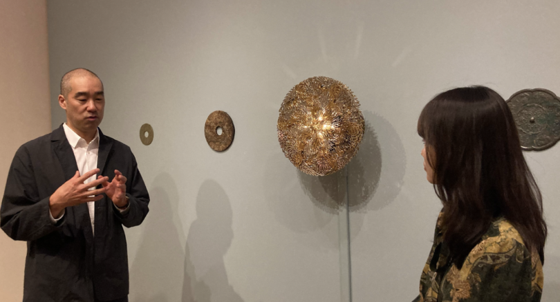 Choe speaks to the left of the Gold Cakra Lamp, a golden disc mounted to the wall, flanked by two metal discs on either side. To the right of the image is Chang, listening to Choe's explanation.