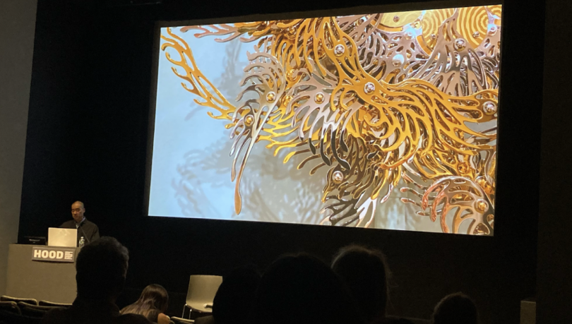 Choe speaking at his artist talk. Choe stands in the bottom left corner of the image, behind a podium. Behind Choe is a large projection of the Gold Cakra Lamps' details.
