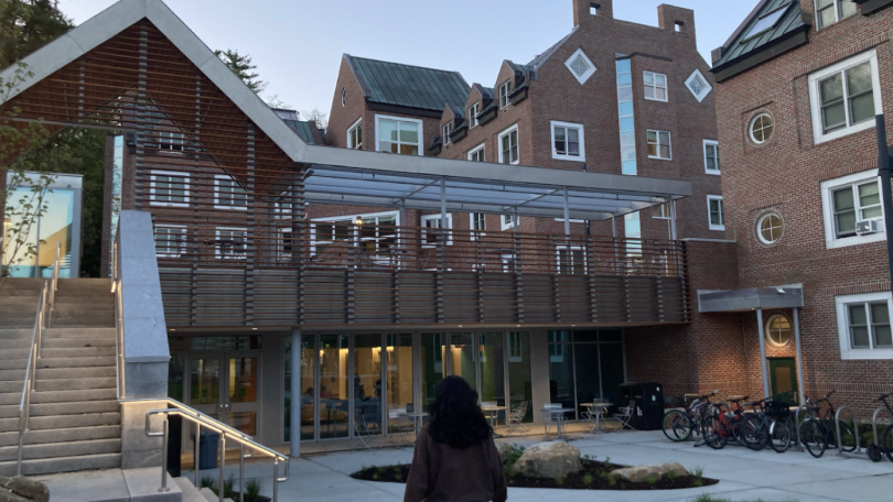 A closer view of Brace Commons. Val is walking, centered in the frame, with her back facing the camera. To her left is a large staircase leading up to the rooftop. In the middle of the image is the entrance to Brace Commons, and to the right is Morton Hal