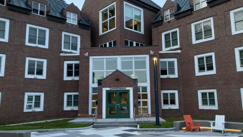 The front entrance of Zimmerman Hall. The entrance is set on a diagonal in the corner of the L-shaped building. The building is red brick, decorated with large square windows with white trim. To the right of the image are two deck chairs, one orange and o