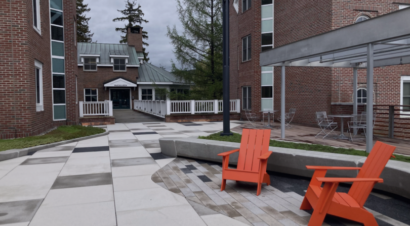 A view of the right of the Brace Commons rooftop. The rooftop is laid with concrete tiles in various shades of grey. To the right of the image are two bright orange lawn chairs. Behind the chairs are sets of silver tables and chairs. In the distance is a