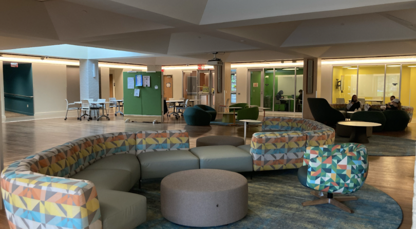 A view of the inside of Brace Commons. The center is an open space containing colorful couches and chairs. A few people are seated in a circle of chairs on the left. In the distant left are two study rooms, with green and yellow walls respectively.