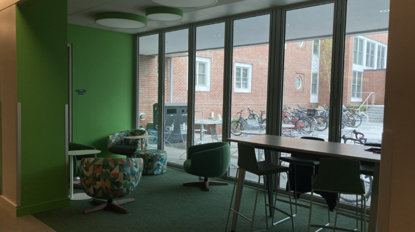 A collection of chairs and tables sit in a green-carpeted area. To the right of the image is a wall of floor-to-ceiling windows, looking out to Morton Hall and the Brace Commons lawn.