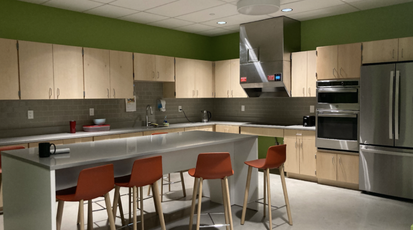 A view of the Brace Commons Kitchen. Light-colored wooden cabinets line the walls. To the right of the room are shiny silver appliances: a stovetop, a fume hood, a microwave on top of an oven, and a double-door refrigerator. In the center of the room is