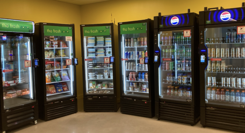 A view of the Brace Commons Fresh Zone, with six refrigerators lined up against a yellow-colored wall. The refrigerators have glass doors, displaying the various snacks and drinks they contain.