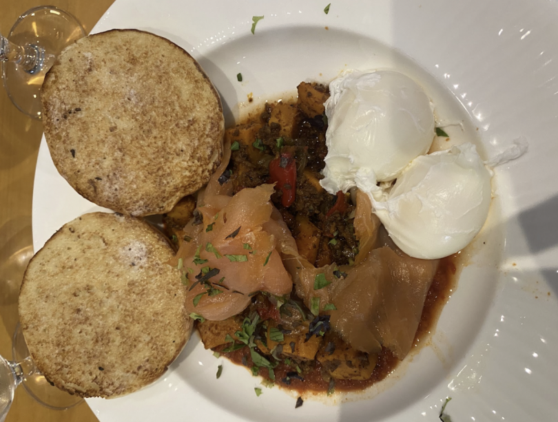 A brunch plate with poached eggs, smoked salmon, vegetable hash, tomato sauce, and two toasted English muffin halves.