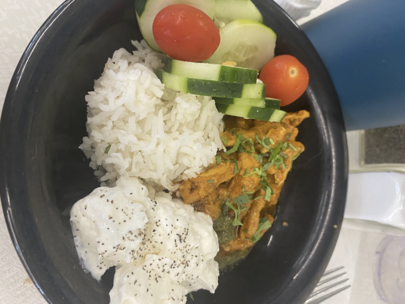 A rice bowl with white rice, creamy yogurt with pepper, cucumber and tomato slices, and a flavorful chicken curry topped with herbs.