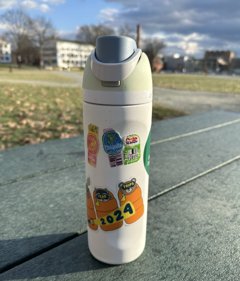 An image of a white, green, and blue Owala brand metal water bottle on a dark green table. The bottle has various stickers covering the outside, including one large one that says "2024" as well as three smaller produce stickers. The background is of a lar