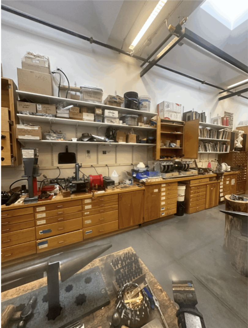 An image of the jewelry studio's cabinet system. In the foreground is a jewelry-forming cone while the central focus of the image is of various brown wooden cabinets and shelves containing various jewelry making tools and materials.