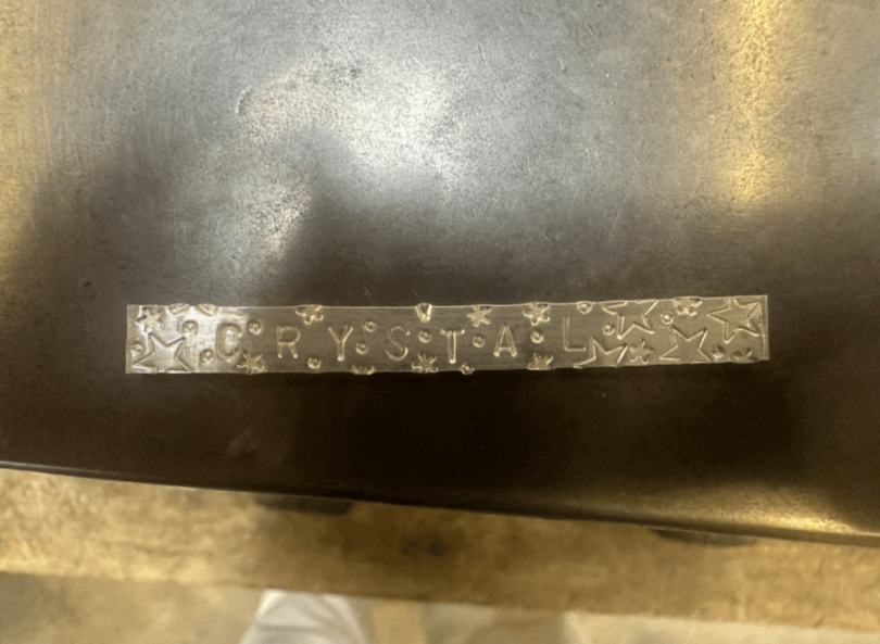 An image of a small silver strip of metal on a black table. The name "Crystal" is etched into the metal while a decorative background of stars and dots are present on the sides.