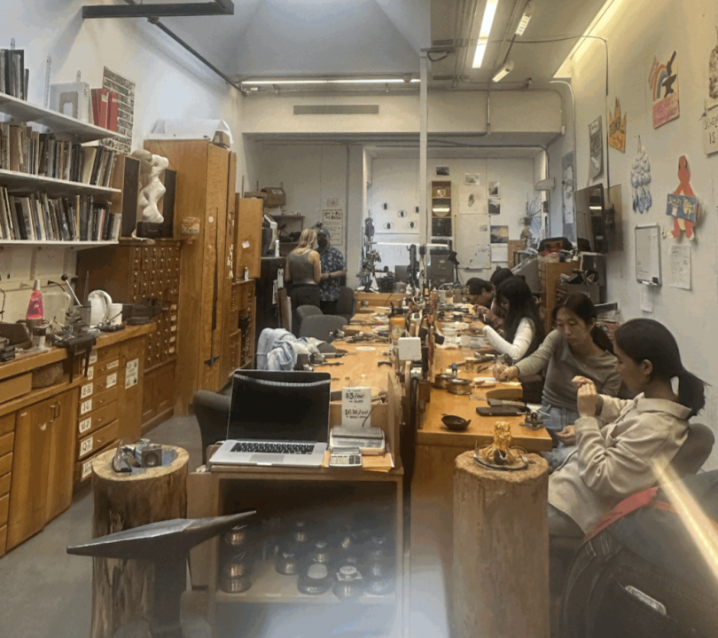 An image of the jewelry studio. There are two long tables with students working at them and cabinets for material storage on the left side of the room. There are multiple students present, all in various stages of working on their projects.