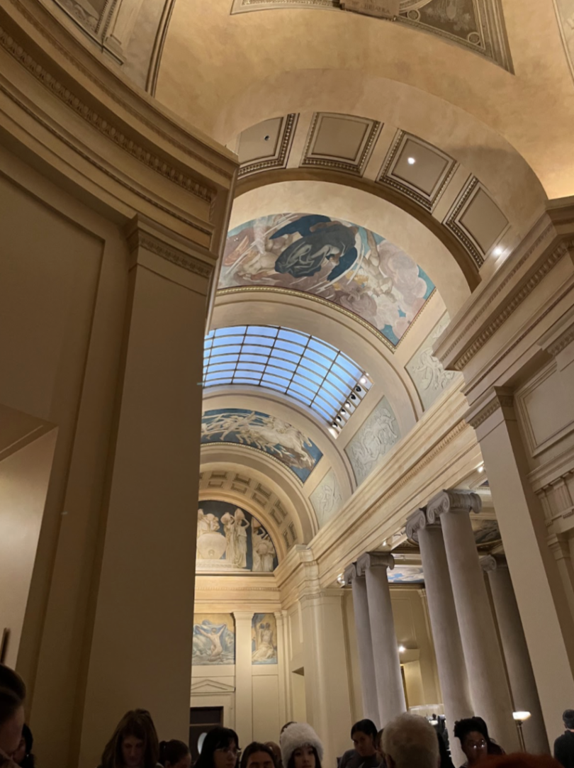 A vertical photo with the camera tilted up to show a beige-colored arch with designs on it. Columns lead down from the arch to the ground where there are people congregating.
