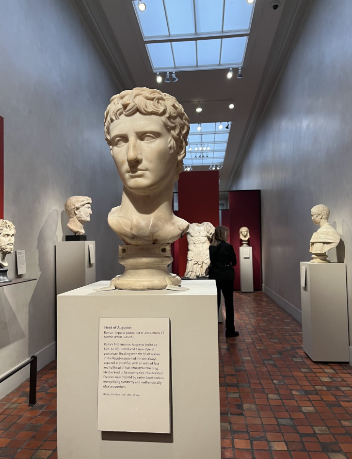 This picture shows a hallway with red and grey tiles on the floor and some windows on the top. There is a marble bust on top of a white pillar in the center o the hallway.