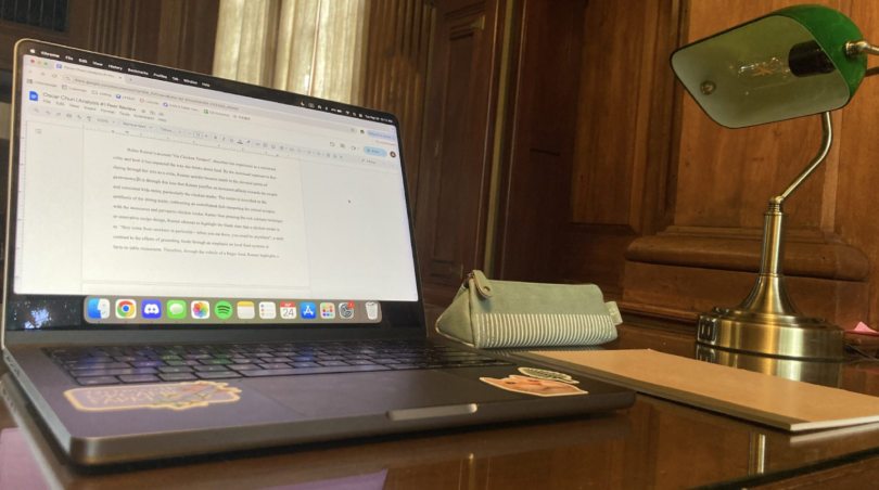 A laptop with a draft from my writing 5 analysis essay, a green pencil pouch, a green lamp, and a brown notebook on a wooden table. The background is a wooden wall and a window covered in a sheer white curtain.