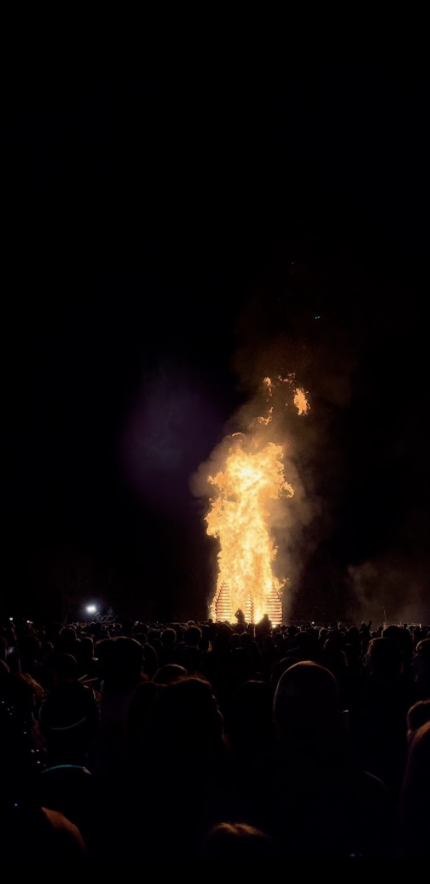 the bonfire glows bright orange and yellow while the crowd of people and night sky behind it are shrouded in darkness.