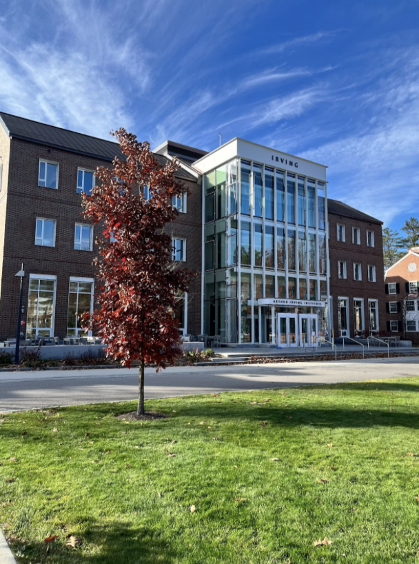 The Irving Institute takes up most of the frame. It's a large building with two brownstone wings and in the middle, where the entrance is, it is glass and white. In the foreground is a tree with red and brown leaves and also there is a small lawn.