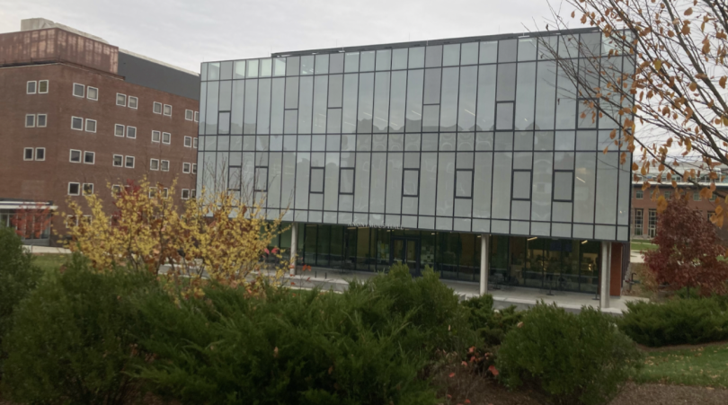 Anonymous Hall, a rectangular glass building, is the the background, with bushes and autumn leaves in the foreground