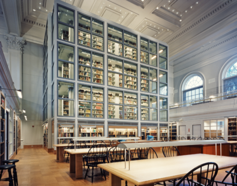 a library with books behind a glass wall surrounded by tables.