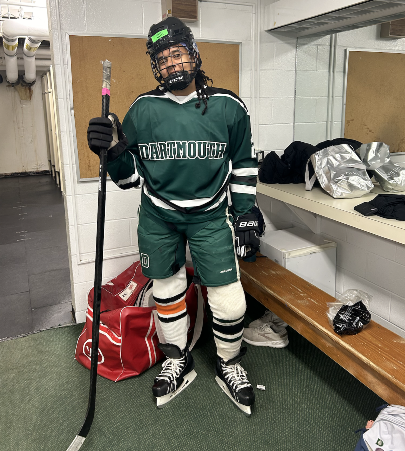 A girl is wearing hockey gear. Her jersey and hockey pants are forest green, bearing the Dartmouth name and logo, while her hockey socks are white. She is wearing skates a helmet, and holds a stick.