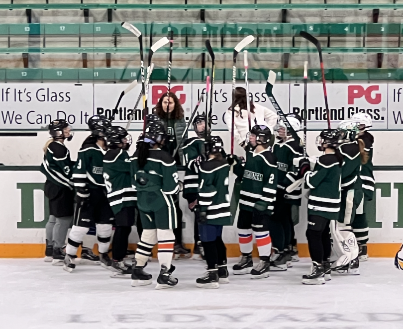 A group of fifteen girls are on the ice in hockey gear. They are huddled together in a circle along with their coach on the bench. All of the girls are holding their hockey sticks up in the air in a cheer.