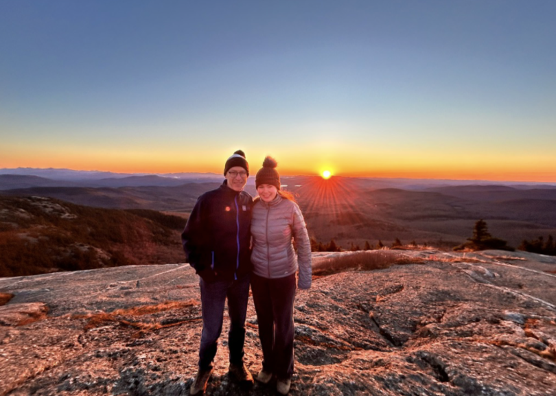 two people at the top of a mountain, the sun is rising behind them