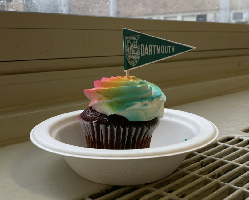 A chocolate cupcake with rainbow-colored frosting with a paper Dartmouth flag stuck in it. The cupcake is in a bowl sitting on a radiator in my high school.
