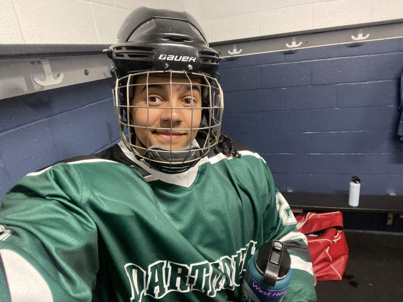 A girl is taking a selfie. She wears a black helmet and a green hockey jersey that reads "Dartmouth." The walls behind her are blue. She is in a locker room.