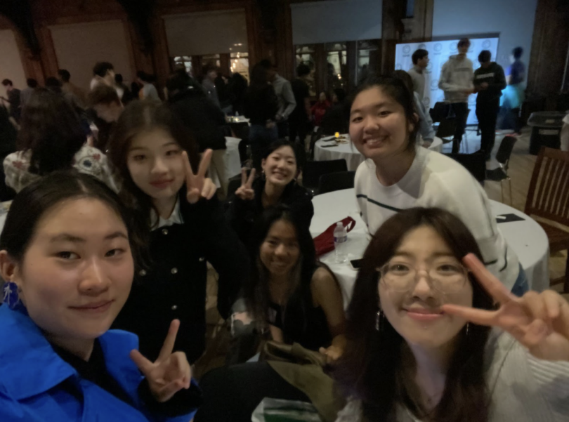 A group of six friends posing together at an ISPOP event. They are smiling and making peace signs with their fingers. The background shows a lively event space filled with other attendees, round tables, and people socializing. The lighting is dim