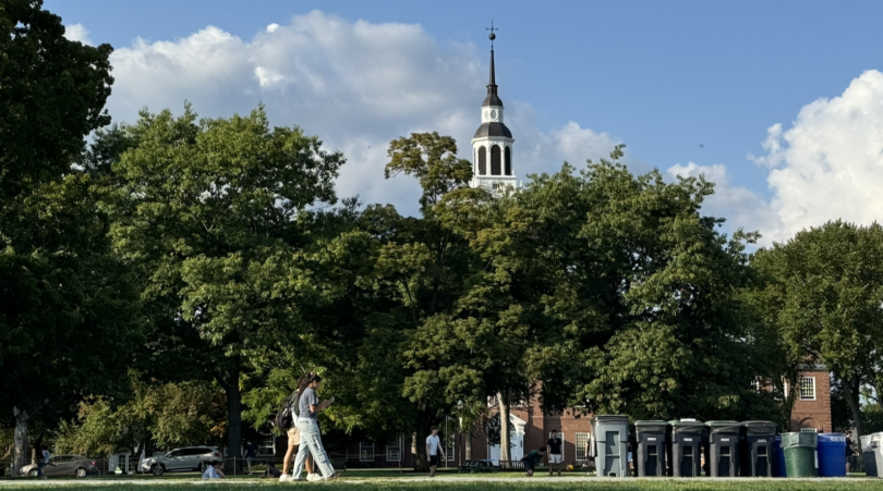 A scenic view of Dartmouth's iconic Baker-Berry Library bell tower rising above a canopy of lush green trees. The sky is bright blue with fluffy white clouds scattered in the background. In the foreground, students are walking across the Green.