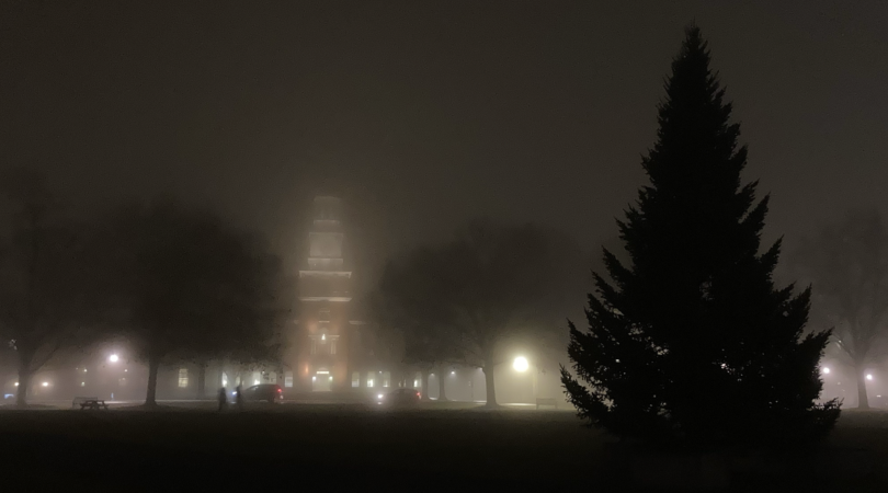 A view of Baker Berry library from the green. The weather is foggy and the sky is dark. In the foreground is the silhouette of a pine tree, in the distance is Baker Berry library. 