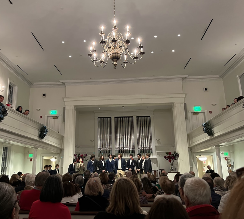 A photo of the inside of a church with white walls with a chandelier hanging from center of the ceiling. Pews line the sides. The backs of people's heads are shown on the bottom of the image. The Brovertones are standing on the center of the stage in form