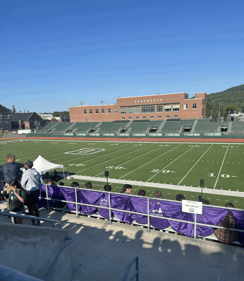 An image taken from the perspective of a spectator in the stands at Dartmouth's Memorial Field. There are students sitting in the stands in front of a walkway. The field is present as the central focus and the stands on the opposite side are visible.