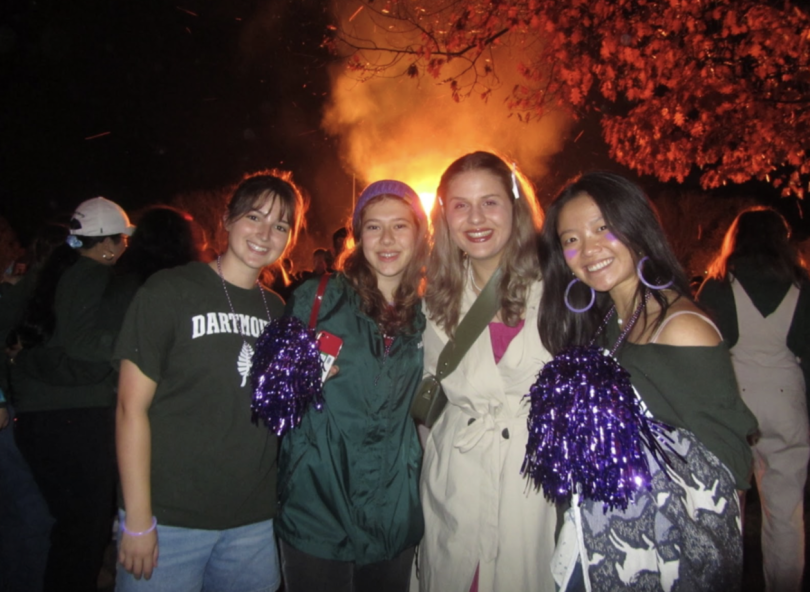 An image of four students in front of a bonfire. The image is taken at night. The students are wearing Dartmouth merchandise and holding purple pom poms.