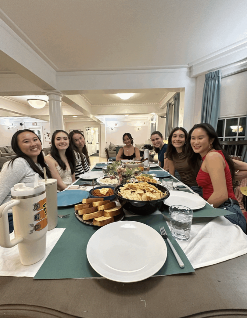 An image of a long dinner table with food and plates. The table is surrounded by students and the background is a well-lit home.