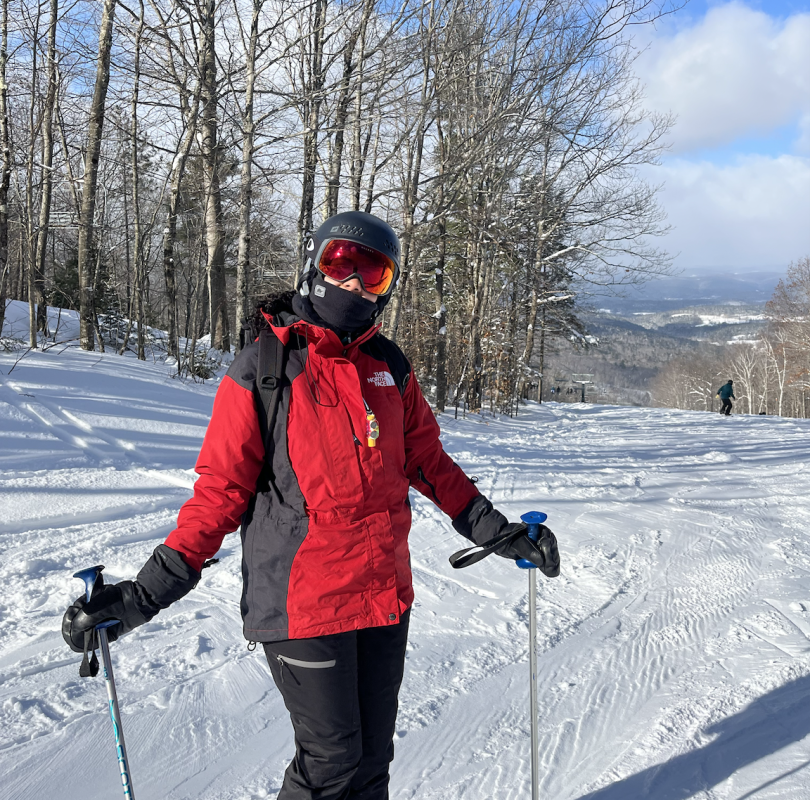 A girl is standing on a ski hill with brown trees behind her on white snow. She is wearing a black helmet, orange/red goggles, a red jacket, and black pants. She is holding two ski poles, one in each hand.