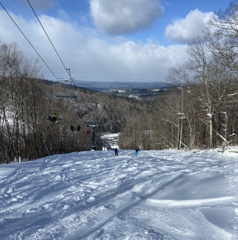 A white ski trail with brown, snow-capped trees lining each side. On the left side is a ski lift with people occupying each seat. People are skiing down the hill, and you can see snowy hills in the background.