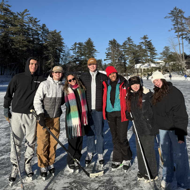 Seven people are lined up, posing for a phone while wearing skates on the ice. Three of them are holding hockey sticks. They all wear colorful clothing and are leaning on one another to squish in for a photo.