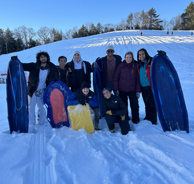 8 people are posing at the base of a snowy hill. 6 are lined up standing, and the 2 on each edge hold long blue sleds. 2 people are kneeling in front of the group. There is a red and blue sled and a yellow sled next to the people in the front.