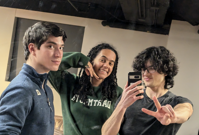 three people are in front of a mirror, taking a photo. The man on the left is standing to the side, a girl in the center is wearing a Dartmouth sweater & posing with her hand under her chin. The man taking the photo is on the right with a peace sign.
