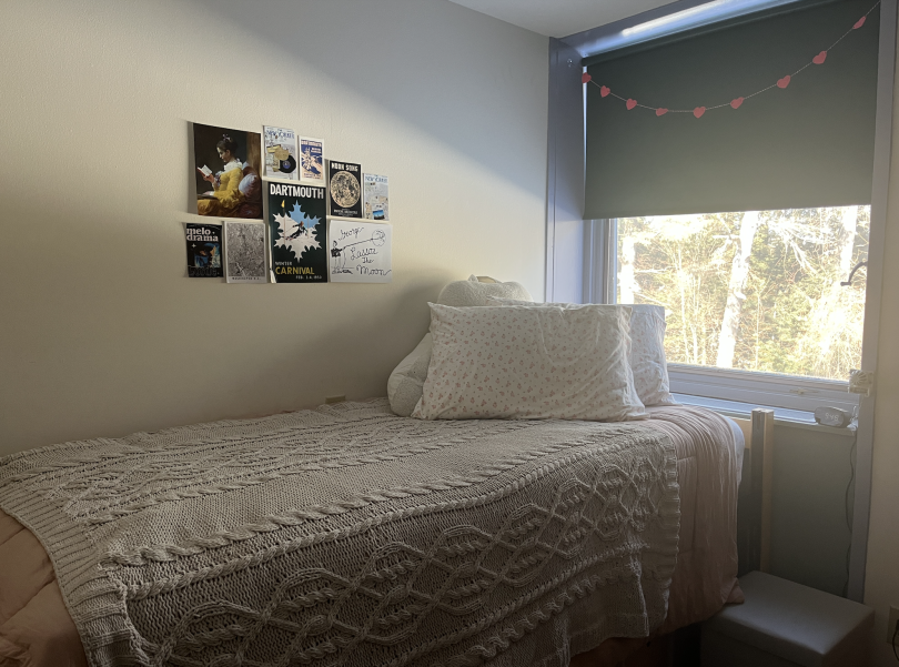 A room with white walls and a window with a green shade. A bed is in the center of the room with a pink comforter and pillows. The wall has posters hanging on it.