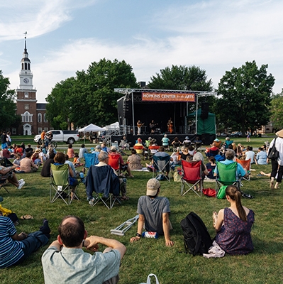 A photo of a concert taking place on the Green with people sitting on the grass and Baker Library in the background