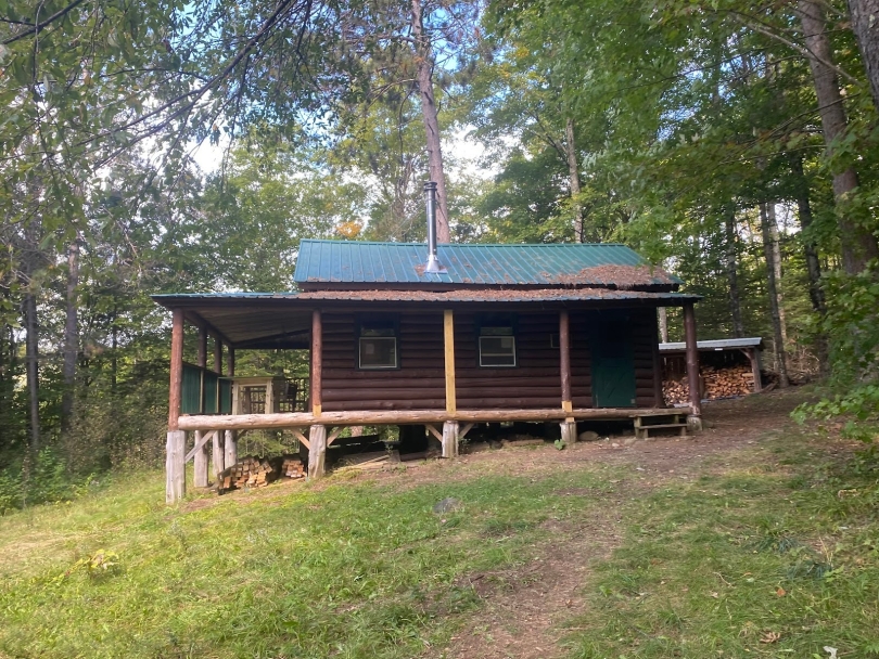 A wooden cabin with a green roof and two windows sitting in the middle of a grassy field. In the front of the building there is a porch, and in the background there is chopped wood for making fires.